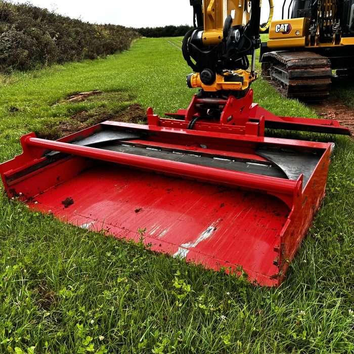 A close-up of a red Turf Translocation Bucket attachment in the foreground with a yellow Caterpillar excavator in the background, set against a rural landscape under a cloudy sky.
