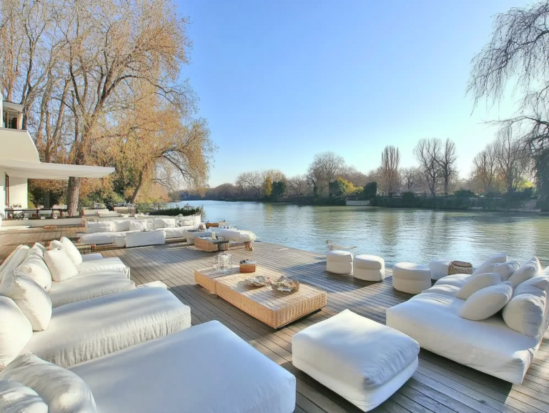 A spacious wooden deck overlooking a calm river, furnished with large white sofas and surrounded by tall trees under a clear sky