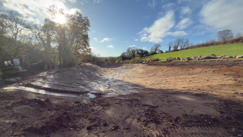 Empty wildlife pool with its liner backfilled, showing excavated granite rocks stacked at the side ready for placement